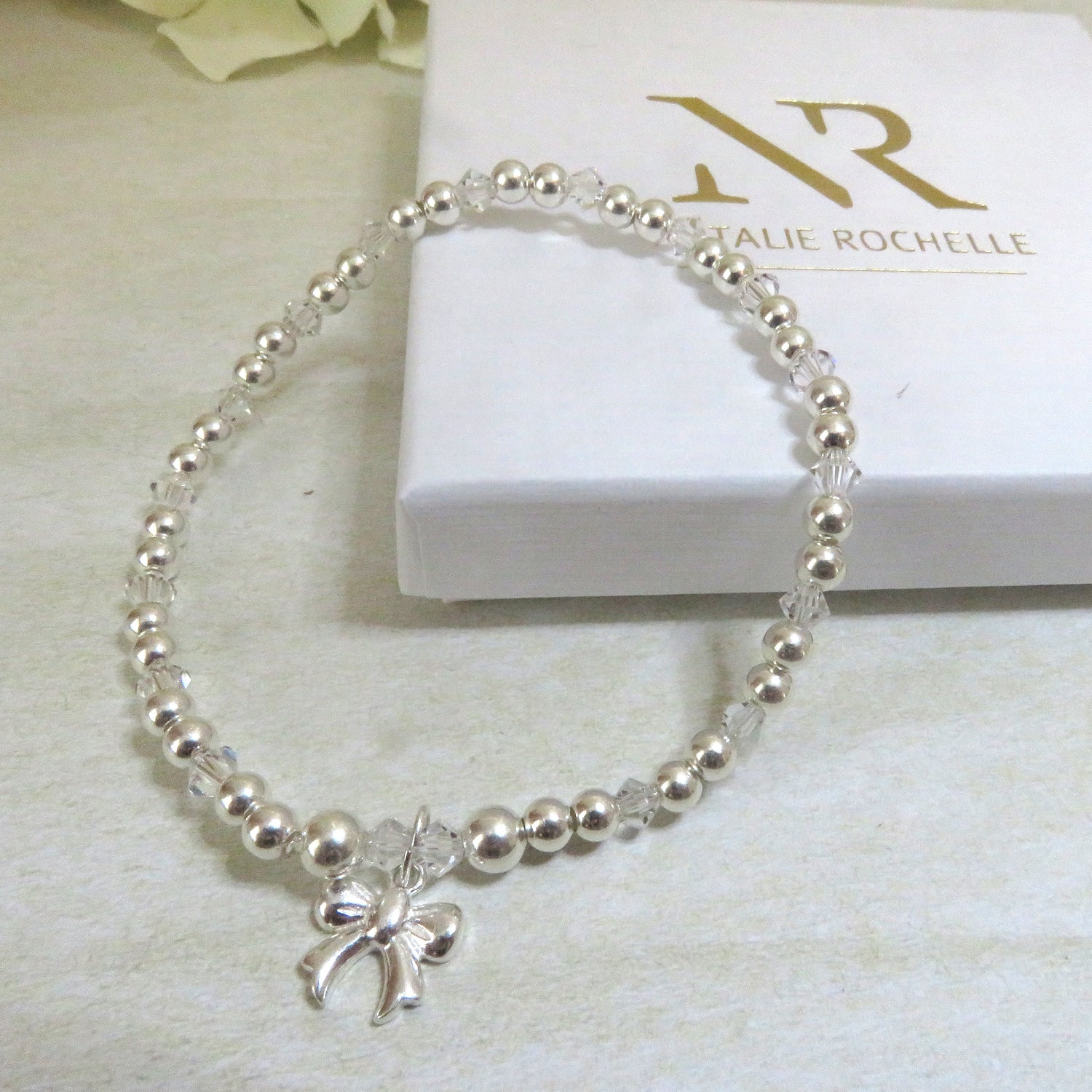 A silver bracelet with crystal beads and a bow charm, displayed on a white box with 'Natalie ROCHELLE' branding.